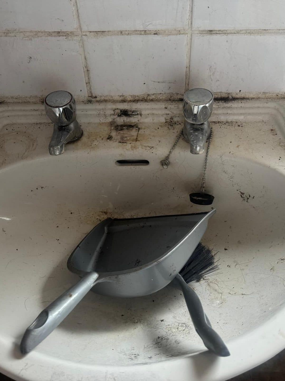 Basin with grime around the taps and a dustpan left in the bowl